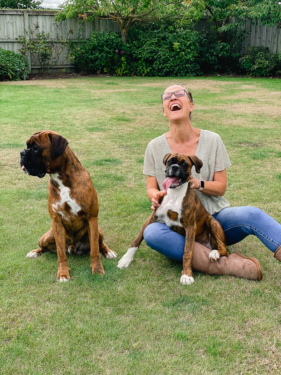 Sharyn laughing on the lawn with her dog Toby in her lap and her dog Floyd to the left not looking at the camera